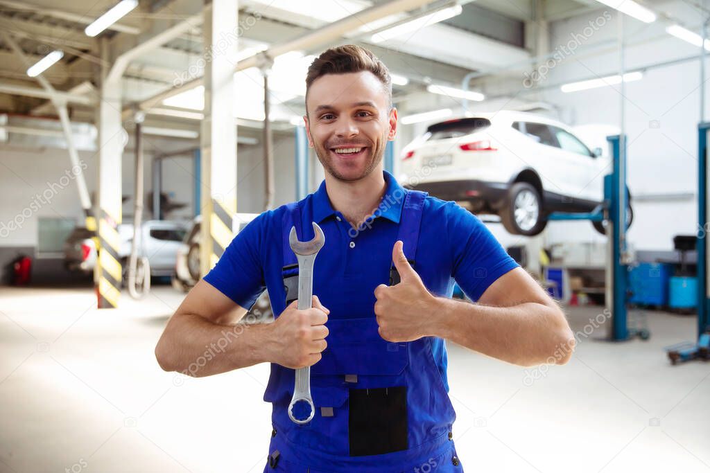 Confident handsome young and experienced car repair worker in work overalls posing against the background of lifted cars in a car service