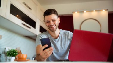 Dizüstü bilgisayarda çalışan ve evde, mutfakta akıllı telefon kullanan mutlu ve serbest çalışan genç adam.