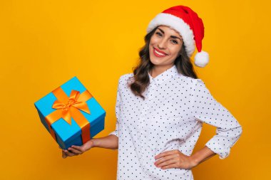 Elinde hediye kutusu olan kırmızı Noel Baba şapkalı güzel bir kadın Noel ve yeni yılı kutlarken izole edilmiş sarı arka planda poz veriyor.