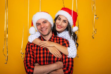 Noel Baba şapkalı mutlu ve heyecanlı genç bir çiftin Noel ve yeni yılı kutlarken dekoratif sarı bir arka plana poz verirken yakın plan fotoğrafı.