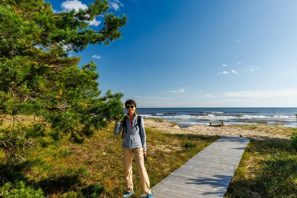 Güneşli bir günde, deniz kenarında tahta kaldırımda yürüyen bir gezgin. Çam ağaçları, kum ve mavi gökyüzü ile kıyı manzarası özgürlüğü, keşfi ve açık hava macerasını simgeliyor.