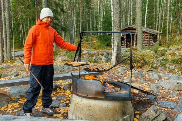 Açık havada giyinmiş biri ormanda kamp ateşinin üstünde yemek pişiriyor. Düşen yapraklarla dolu bir sonbahar sahnesi, arka planda bir barınak ve doğa yaşamını sembolize eden huzurlu bir vahşi yaşam ortamı.