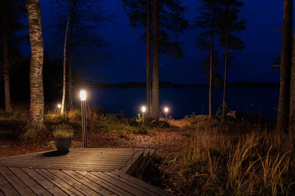 Gece manzarası, sakin bir göle giden ahşap bir iskele, ağaçlar arasındaki ılık bahçe ışıklarının hafifçe aydınlattığı, doğada huzurlu ve rahat bir akşam atmosferi yaratan.