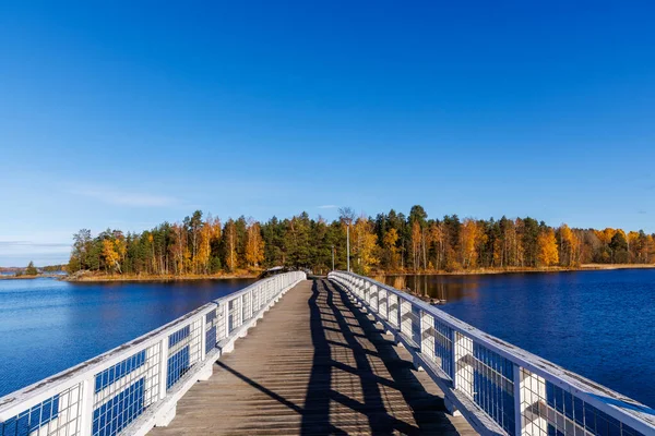 Açık mavi gökyüzünün altında renkli sonbahar ağaçları olan bir adaya doğru mavi bir gölden geçen beyaz tahta bir yaya köprüsünün manzarası