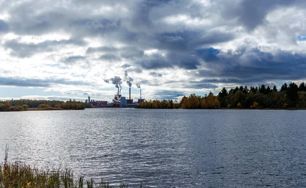 Endüstriyel kağıt fabrikası ya da göl ya da nehir kıyısında bir fabrika dramatik, yoğun bulutlu bir gökyüzü altında, kıyı şeridi boyunca sonbahar yaprakları ile