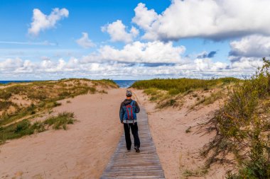 Erkek yürüyüşçü, kum tepelerinden kumsala doğru yürüyor kümülüs bulutlarıyla mavi gökyüzünün altında, huzurlu kıyı manzarası, seyahat, doğa ve açık hava macera projeleri için mükemmel.