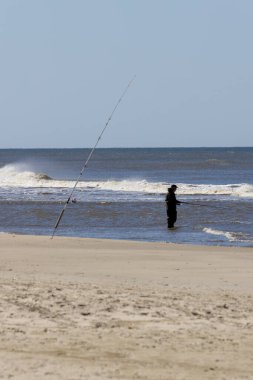 Balıkçı kıyafeti giyen bir adam Danimarka kıyısı boyunca balık tutarken denizde duruyor. Yakınlardaki kumlu plaja bir olta konur. Görüntü Mayıs 2025 'te çekildi ve sakin bir deniz balıkçılığı ve açık hava eğlencesi anını yansıtıyor.