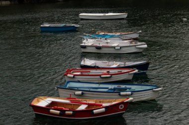 Derin ve karanlık sularda yüzen birkaç parlak renkli ahşap teknenin yüksek açılı görüntüsü. Yüzeyin durgunluğu aynaya benzer bir etki yaratıyor, boyanın canlı renklerini ve sakin bir limanda demirleyen gemilerin yalnızlığını vurguluyor.