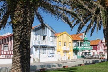 Costa Nova, Aveiro 'da, Palheiros olarak bilinen ikonik geleneksel ahşap evlerin yer aldığı canlı ve neşeli bir sokak sahnesi. Görüntüde mavi, sarı ve yeşil çizgileri olan büyüleyici binalar sergileniyor.