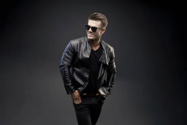 handsome young man in sunglasses and leather jacket with glasses and bag on black background