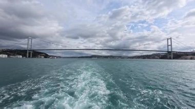 İstanbul, Türkiye 'de boğaza yayılan Boğaz Köprüsü dramatik bulutlar ve turkuaz suyu ile feribottan görülüyor