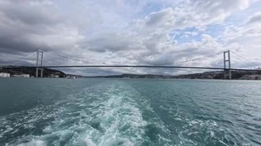 İstanbul, Türkiye 'de boğaza yayılan Boğaz Köprüsü dramatik bulutlar ve turkuaz suyu ile feribottan görülüyor