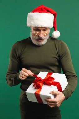 Mutlu sakallı, modern Noel Baba hediyesini tutuyor ve yeşil arka planda şakalaşıyor.