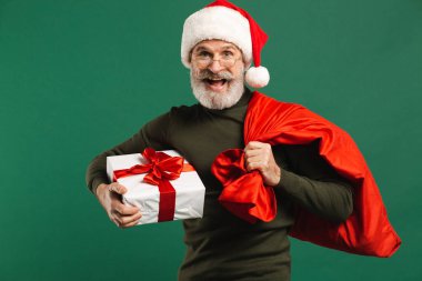 Mutlu sakallı, modern Noel Baba elinde hediye kutusu ve yeşil arka planda kırmızı çanta.