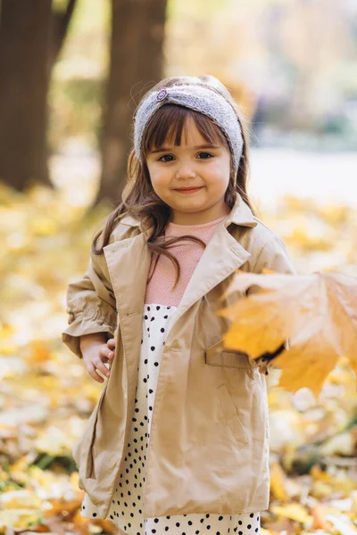 秋天的公园里 穿着米黄色外套 手里拿着一片黄色枫叶的快乐而美丽的小女孩 — 图库照片