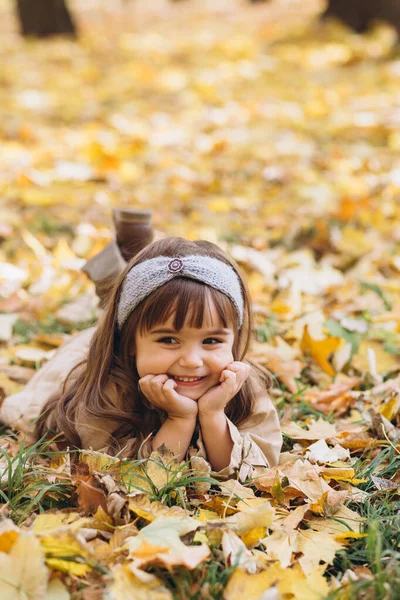 秋天公园的黄叶中 躺着一位穿着米黄色外套的美丽快乐的小女孩 — 图库照片