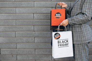 Gri takım elbiseli kadın, gri tuğla duvarlı arka planda kağıt torbalar tutuyor.