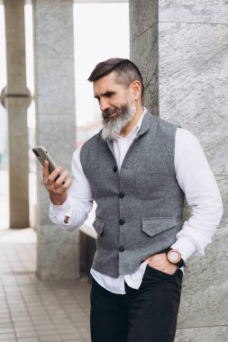 Sakallı, kır saçlı, yaşlı bir adam akıllı telefon kullanır ve konuşur.