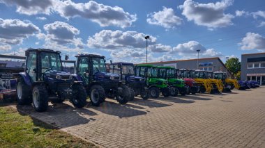 Winsen, Almanya; 30 Eylül 2025: Yeni Farmtrac ve Deutz-Fahr traktörleri mavi gökyüzünün altındaki ithalat sahasına park edilmiş, mavi, yeşil ve kırmızı olmak üzere çeşitli renkler müşteriler için modern kompakt tarım makinelerini temsil etmektedir..