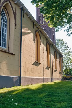 Usquert, Hollanda; 11 Haziran 2023: Kemerli camlı eski kilisenin sarı alçı duvarı ve yeşil ağaçların arasındaki tuğla kule manzarası.