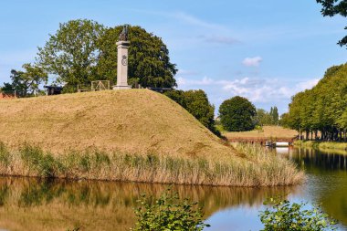 Landskrona, İsveç - 8 Ağustos 2025: Landskrona Kalesi 'ndeki çimenli bir tepenin üzerinde belirgin bir heykel bulunan ve berrak gökyüzü yeşilliğini ve küçük bir köprüyü yansıtan bir hendekle çevrili anıtsal bir sütun duruyor..