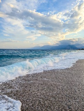 Gentle Mediterranean waves roll onto pebble beach under dramatic cloudy sky during golden hour near Antalya Turkey coast. High quality photograph