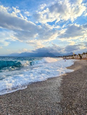 Akdeniz dalgaları, Antalya Türkiye kıyı şeridi yakınlarında dramatik günbatımı gökyüzü altında beyaz köpük oluşturan kayalık çakıl taşlı plaja çarpar. Yüksek kalite fotoğraf