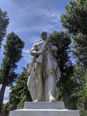 White stone Madonna statue with praying hands stands peacefully among green trees at outdoor shrine in northern Belarus. High quality photograph.