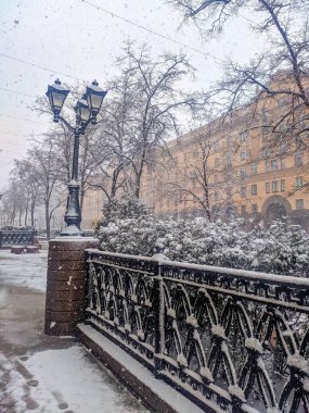 Minsk, Belarus, May 01, 2025. Snowy winter scene with ornate metal fence covered in fresh snow and historic building visible through bare trees. High quality photograph