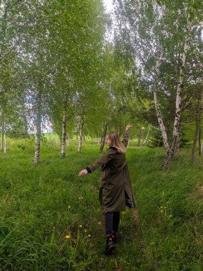 Novorudok, Belarus, May 01, 2025. Young woman in olive jacket enjoying spring day among vibrant green birch trees in peaceful forest setting. High quality photograph
