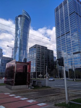 Warsaw, Poland, May 01, 2025. Modern commercial district with glass office towers and pedestrian walkway featuring colorful red pavement under blue cloudy sky. High quality photograph