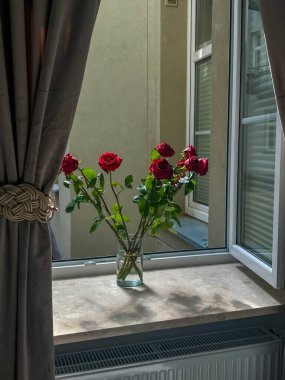 Varşova, Polonya, Mayıs 01, 2025. Cam vazo, pencerenin kenarında duran taze kırmızı güller ve yumuşak doğal gün ışığı kapalı alanda romantik bir atmosfer yaratıyor. Yüksek kalite fotoğraf