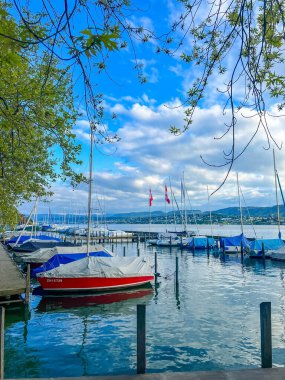 Zürih, İsviçre, Mayıs 01, 2025. Zürih Gölü 'nün kırmızı ve mavi tekneleri dramatik bulutlu gökyüzünün altında limana demirlemiş canlı deniz manzarası yaratıyor. Yüksek kalite fotoğraf