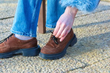 Varşova, Polonya, Mart 01, 2026. Taştan bir kaldırımda mavi kot pantolon giymiş kahverengi deri botlara dokunan ellerin aksiyon çekimi. Yüksek kalite fotoğraf.