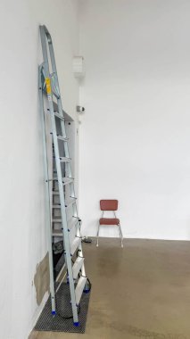Varşova, Polonya, 15 Mart 2026. Boş beyaz bir sanayi stüdyosunda uzun metal bir merdiven ve tek bir kırmızı sandalye. Yüksek kalite fotoğraf.