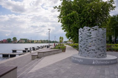 Polonya 'nın Dziwnow kentindeki modern bir rıhtım güvertesi, balık kesimleriyle eşsiz silindirik bir heykel içeriyor. Gezinti güvertesi gri fayanslarla döşelidir ve ahşap banklar, sokak lambaları ve yeşil ağaçlara sahiptir. Sola, sayısız vızıltısı olan bir su kütlesi.