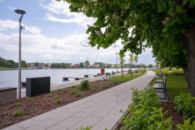 Polonya 'nın Dziwnow kentindeki modern, asfalt bir su kütlesi. Yeşil yaprakları ve beyaz çiçekleri olan kestane ağaçları, banklar ve sokak lambalarının yer aldığı yolun sağ tarafını kaplıyor. Ahşap rıhtımlar soldaki suya doğru uzanıyor.,