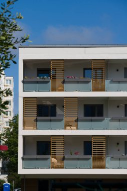 Güneşli bir günde modern bir apartmanın dışında. Ön cephede buzlu cam korkuluklar ve dekoratif ahşap çıtalarla çağdaş balkonlar yer alıyor. Önplandaki yeşil bir ağaç şehir ortamına doğanın bir dokunuşunu ekler. Modern için kavram