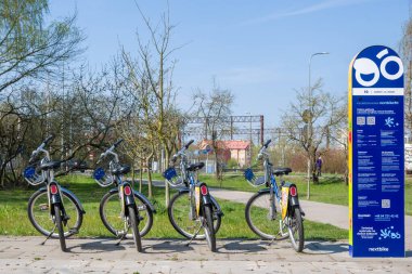 Güneşli bir bahar gününde, bir şehir parkındaki Nextbike kiralama istasyonunda bir sıra bisiklet. Arka planda demiryolu ve ağaçlar var. Bu resim modern şehir hareketliliğini, çevre dostu ulaşımı, sürdürülebilirliği ve bir sağlıkçıyı temsil ediyor.