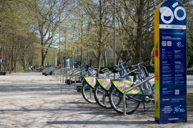 Metal raflarda parketmiş birkaç Nextbike bisikleti olan bir bisiklet paylaşım istasyonu. İstasyon, bir şehir parkında ya da ormanda, taze bahar yapraklı uzun ağaçlarla çevrili bir patikada yer almaktadır. Arka planda ahşap bir bank görünür. Görüntü,