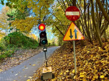 Altın yapraklarla kaplı dar bir patika, kırmızı gösteren geçici trafik ışığı ve parlak sarı ağaçların arasında duran yol işaretleriyle sonbahar parkı sahnesi. Doğa ve şehir elementlerinin çarpıcı kontrastı.