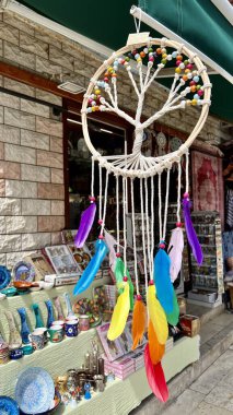 Beyaz Hayat Ağacı desenli, gökkuşağı tüylü ve parlak boncuklu renkli bir düş kapanı seramik ve tekstil ürünleri satan bir hediyelik eşya dükkanının önünde asılı..