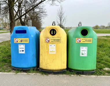 Kağıt, plastik metal ve cam için mavi, sarı ve yeşil renkli üç büyük açık hava geri dönüşüm kutusu, arka planda bulutlu bir gökyüzünün altında çimen ve ağaçlarla birlikte kaldırımda duruyor..