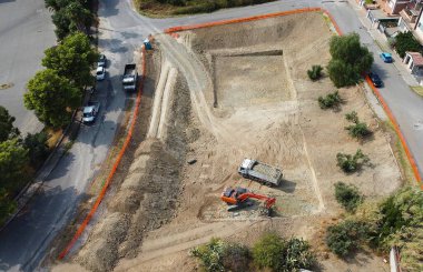 Kazı makinesi ve çöp kamyonu olan bir inşaat alanının havadan görüntüsü. Yer çakıl ve toprakla kaplı, bu da ağır ekipmanların çalışmalarını ve arazi hazırlığının ilk aşamalarını gösteriyor.