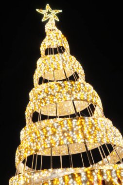 Altın Spiral Noel Ağacı. Karanlık bir gece gökyüzüne karşı, üzerinde parlak bir yıldız bulunan, sıcak beyaz ve altın renkli LED spiralleri olan modern, stilize edilmiş bir Noel ağacının alçak açılı görüntüsü..