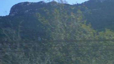 İtalya 'daki bir tren penceresinden panoramik bir orman manzarası ve kayalık tepeler. Yoğun yeşil ormanlar, parlak bir gökyüzünün altındaki dağ yamaçlarını gizler..