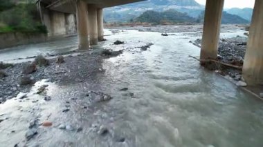 Bir FPV dronu bir dağ nehri üzerindeki köprünün altından uçuyor. Beton barajın, şelalenin ve görkemli kanyonun dinamik manzarası. Doğanın gücü ve kırsal alanlar 4K 'da..