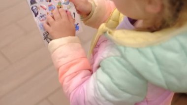 Bir alışveriş merkezinde ailesiyle alışveriş yaparken elinde renkli bir Cadılar Bayramı çıkartması tutan ve denetleyen bir anaokulu kızının yakın çekimi. 