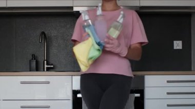 Close-up of a woman in pink gloves holding three spray bottles with colorful liquids and microfiber cloths in a modern kitchen interior. Eco-friendly cleaning concept