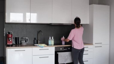 Female in pink T-shirt and rubber gloves wiping a glossy white kitchen cabinet using a blue microfiber cloth. Daily household cleaning routine in a modern apartment interior